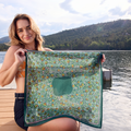 Woman holding a green patterned fabric by a lake with mountains in the background