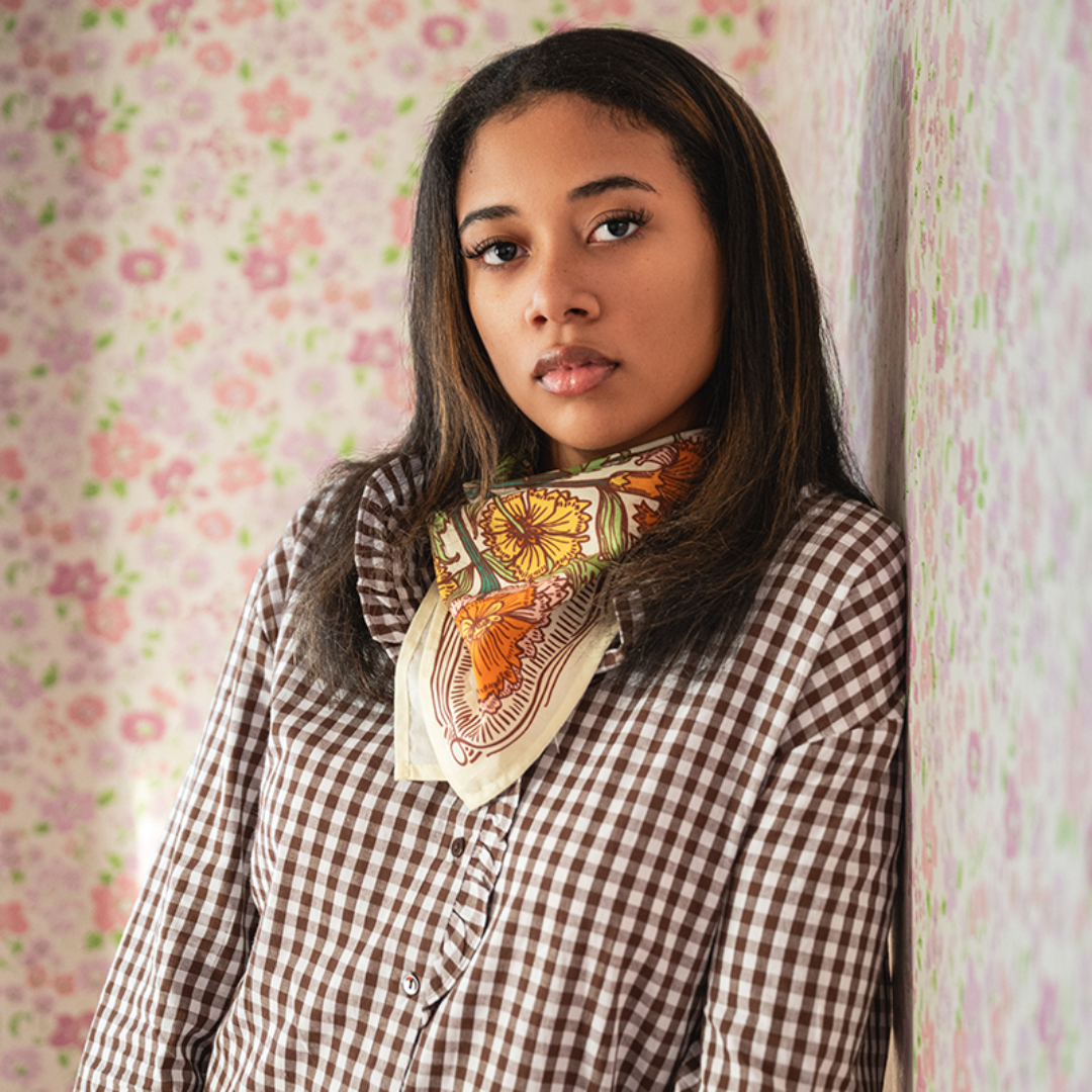 Woman wearing a checkered shirt with a patterned scarf against a floral wallpaper background