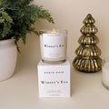 Candle labeled 'Winter's Eve' by North Rose with packaging, potted plant, and decorative tree on a neutral background