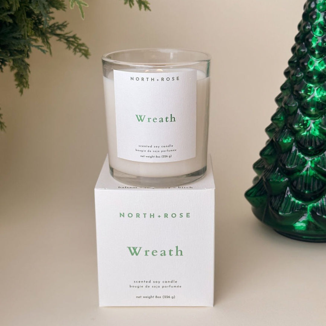 Candle labeled 'Wreath' by North+Rose on a white surface with a green decorative item.