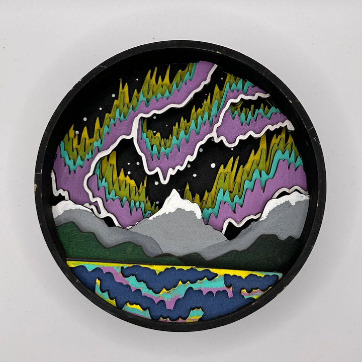 A Paint By Layers Craft Kit featuring a circular shadow box with a pre-designed landscape scene. The scene depicts a mountain range under a vibrant aurora borealis. The kit includes layers of wood, paint, and instructions. The title of the kit is 