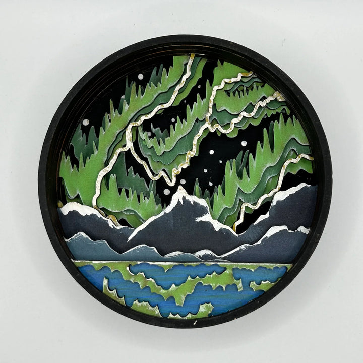 A Paint By Layers Craft Kit featuring a circular shadow box with a pre-designed landscape scene. The scene depicts a mountain range under a vibrant aurora borealis. The kit includes layers of wood, paint, and instructions. The title of the kit is 