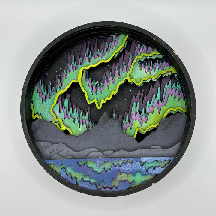 A Paint By Layers Craft Kit featuring a circular shadow box with a pre-designed landscape scene. The scene depicts a mountain range under a vibrant aurora borealis. The kit includes layers of wood, paint, and instructions. The title of the kit is 