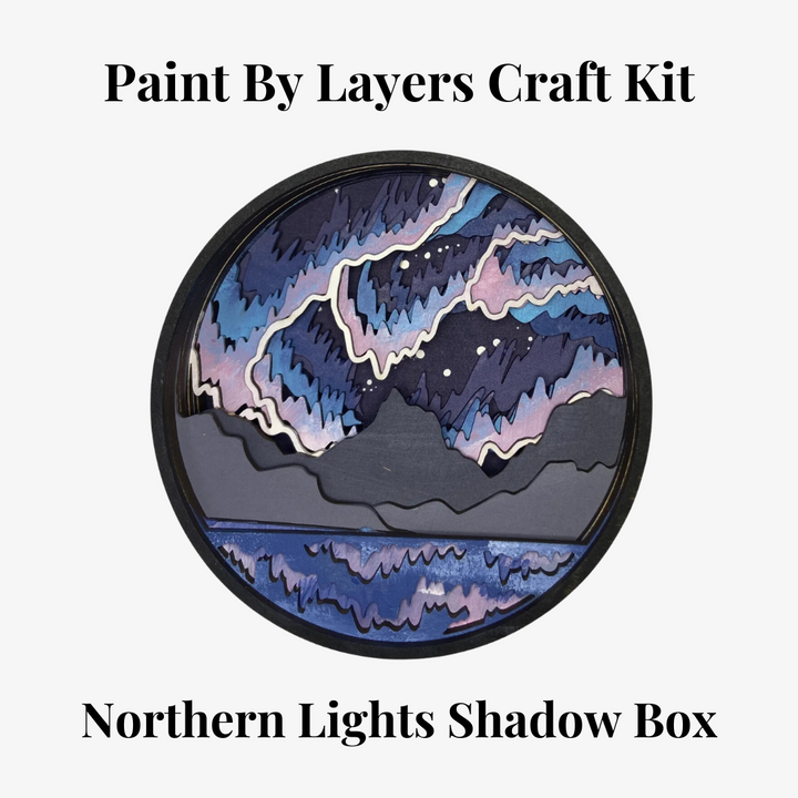 A Paint By Layers Craft Kit featuring a circular shadow box with a pre-designed landscape scene. The scene depicts a mountain range under a vibrant aurora borealis. The kit includes layers of wood, paint, and instructions. The title of the kit is 