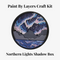 A Paint By Layers Craft Kit featuring a circular shadow box with a pre-designed landscape scene. The scene depicts a mountain range under a vibrant aurora borealis. The kit includes layers of wood, paint, and instructions. The title of the kit is 