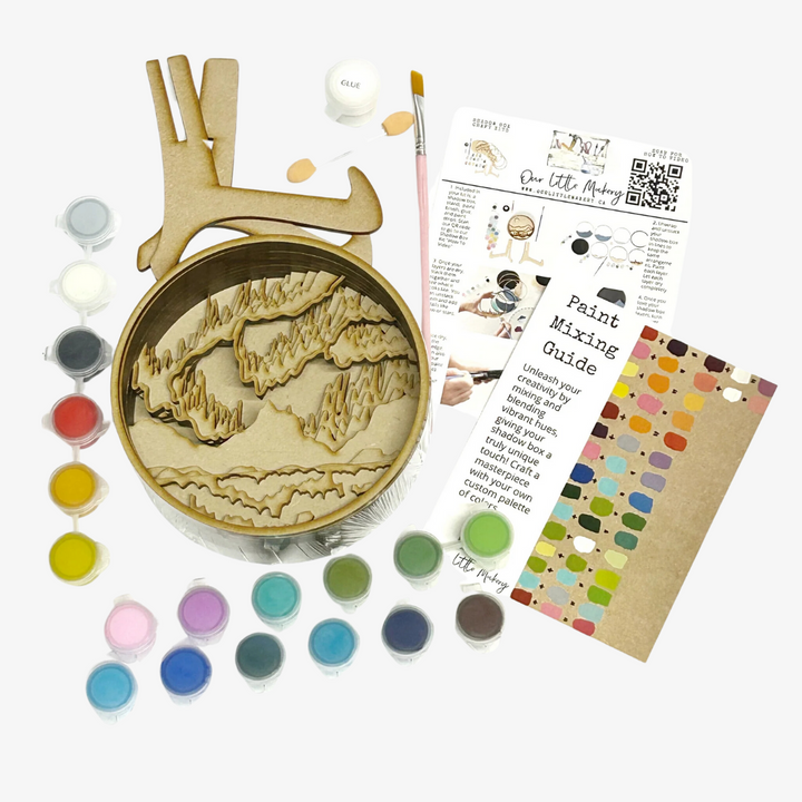 A Paint By Layers Craft Kit featuring a circular shadow box with a pre-designed landscape scene. The scene depicts a mountain range under a vibrant aurora borealis. The kit includes layers of wood, paint, and instructions. The title of the kit is 