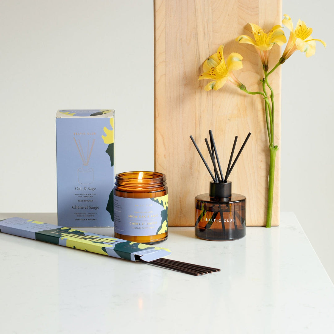 Candle, diffuser, and packaging on a white surface with yellow flowers in the background