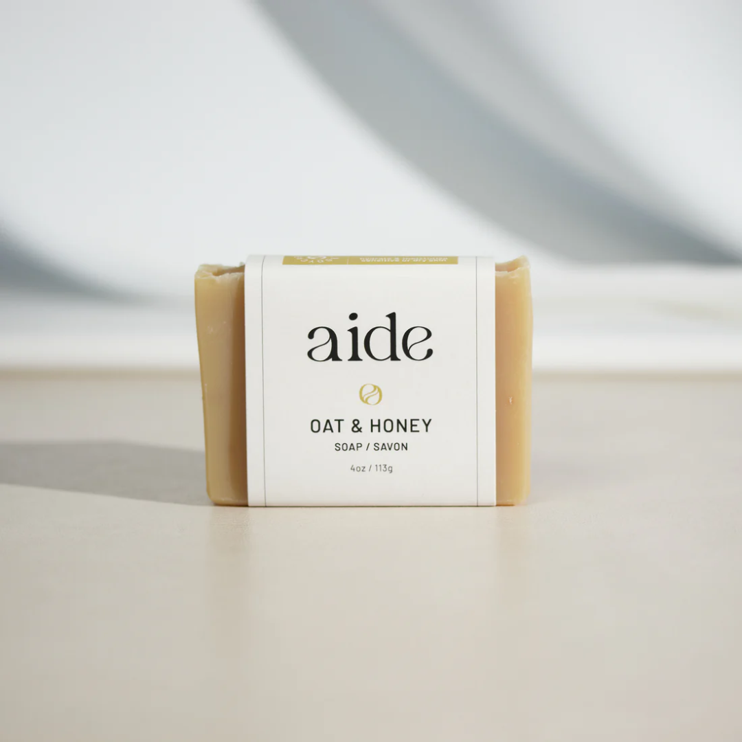 A bar of Aide Bodycare Oat & Honey Soap, featuring a light beige color and a paper label with the 'aide' logo and 'Oat & Honey' text, displayed on a light-colored surface. The soap is natural and handmade, available in Mississippi Mills, Ontario, Canada.