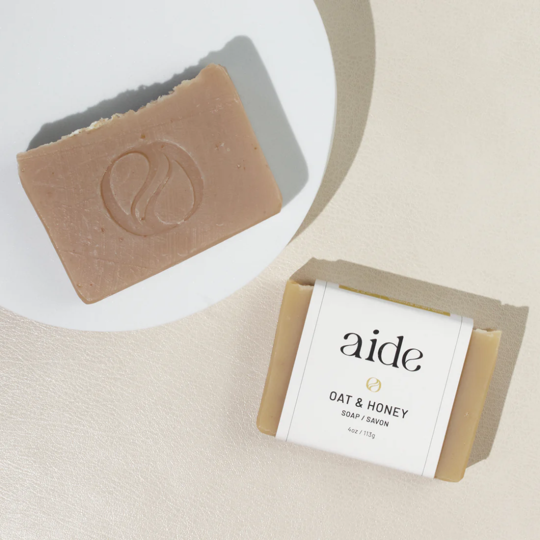 A bar of Aide Bodycare Oat & Honey Soap, featuring a light beige color and a paper label with the 'aide' logo and 'Oat & Honey' text, displayed on a light-colored surface. The soap is natural and handmade, available in Mississippi Mills, Ontario, Canada.