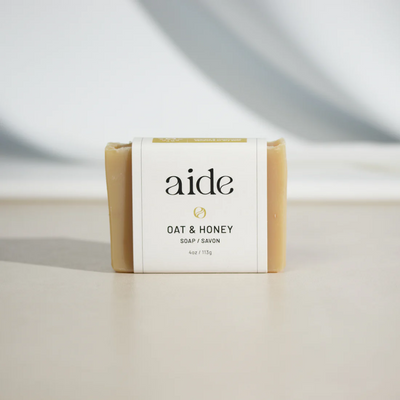 A bar of Aide Bodycare Oat & Honey Soap, featuring a light beige color and a paper label with the 'aide' logo and 'Oat & Honey' text, displayed on a light-colored surface. The soap is natural and handmade, available in Mississippi Mills, Ontario, Canada.