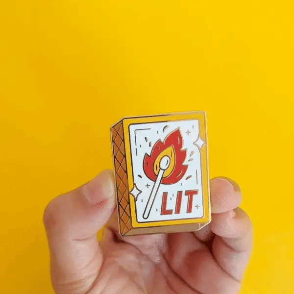 A hand holding a gold-colored enamel pin shaped like a matchbox. The matchbox has a lit match with a flame and the word 