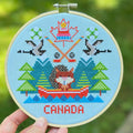 Cross-stitched design of a fox in a canoe with 'Canada' text, held in front of a blurred green background.
