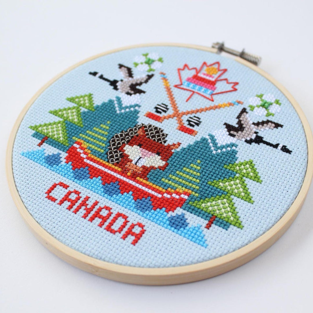 Cross-stitched design of a Canadian flag with trees and a canoe, framed in an embroidery hoop.