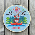 Cross-stitched design of a Canadian-themed scene with trees, a maple leaf, and 'Canada' text on a wooden background.