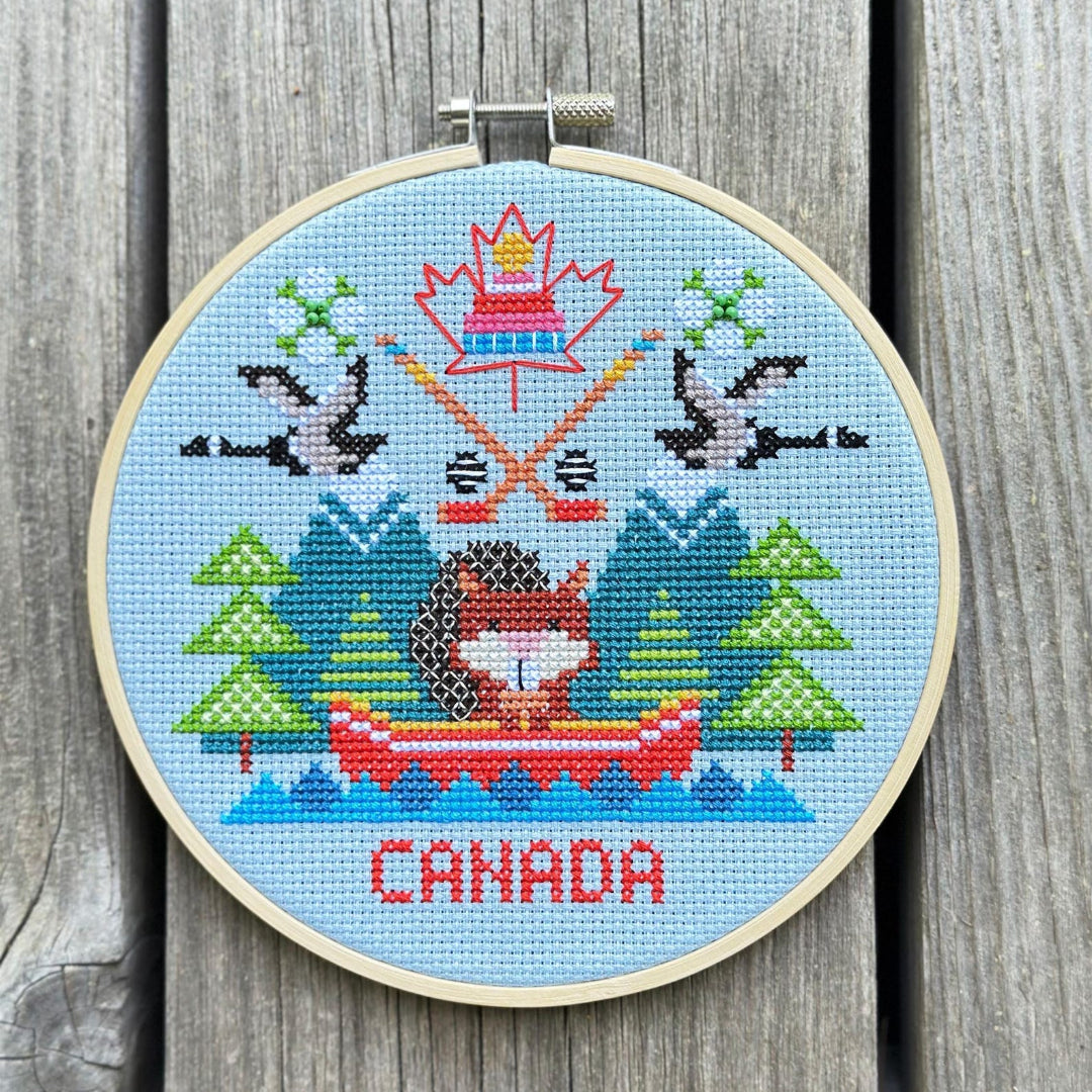 Cross-stitched design of a Canadian-themed scene with trees, a maple leaf, and 'Canada' text on a wooden background.