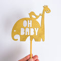 Gold cake topper with elephant, bird, and giraffe shapes and 'OH BABY' text on a white background.