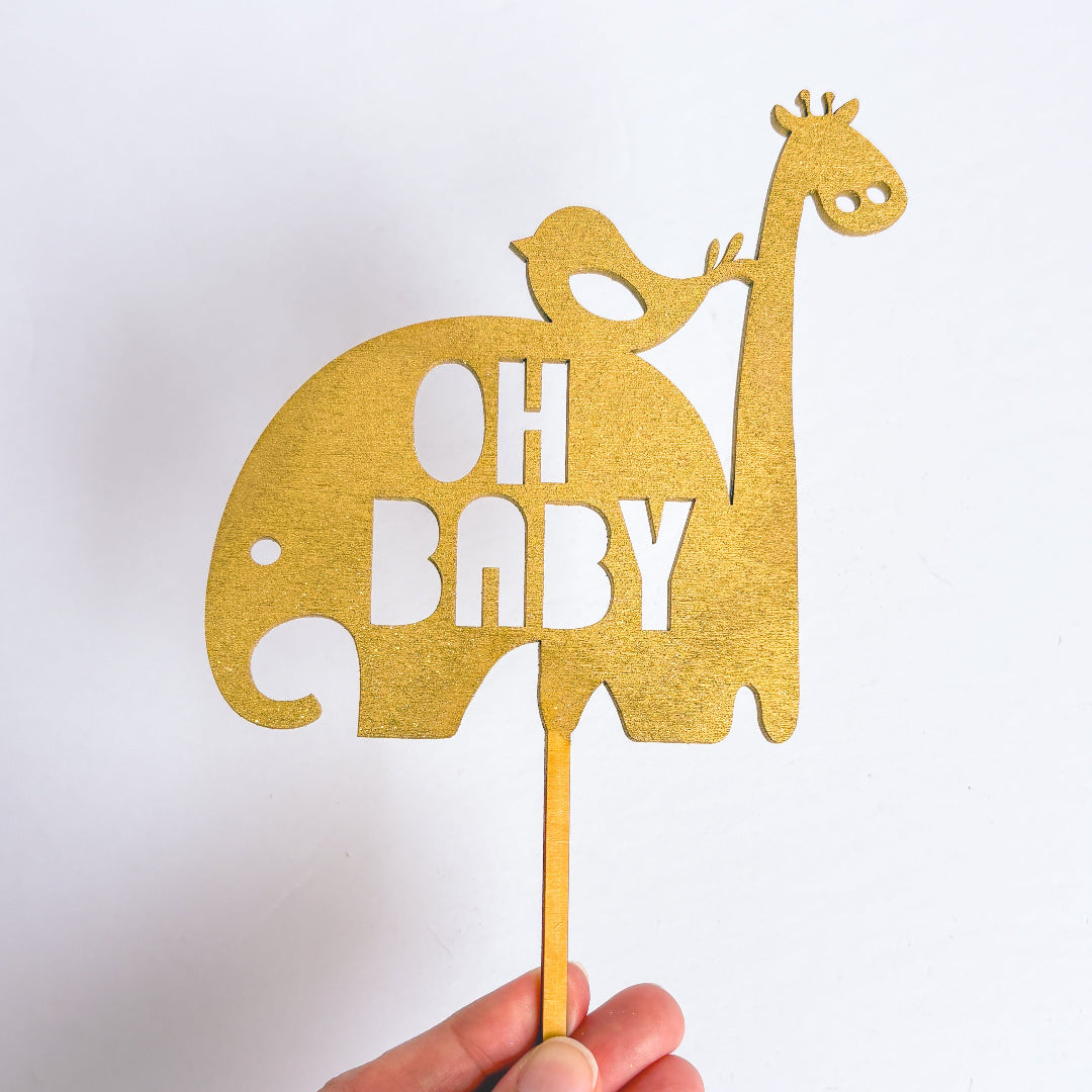 Gold cake topper with elephant, bird, and giraffe shapes and 'OH BABY' text on a white background.