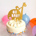Small white cake with colorful sprinkles, a gold 'OH BABY' giraffe topper, and balloons in the background.