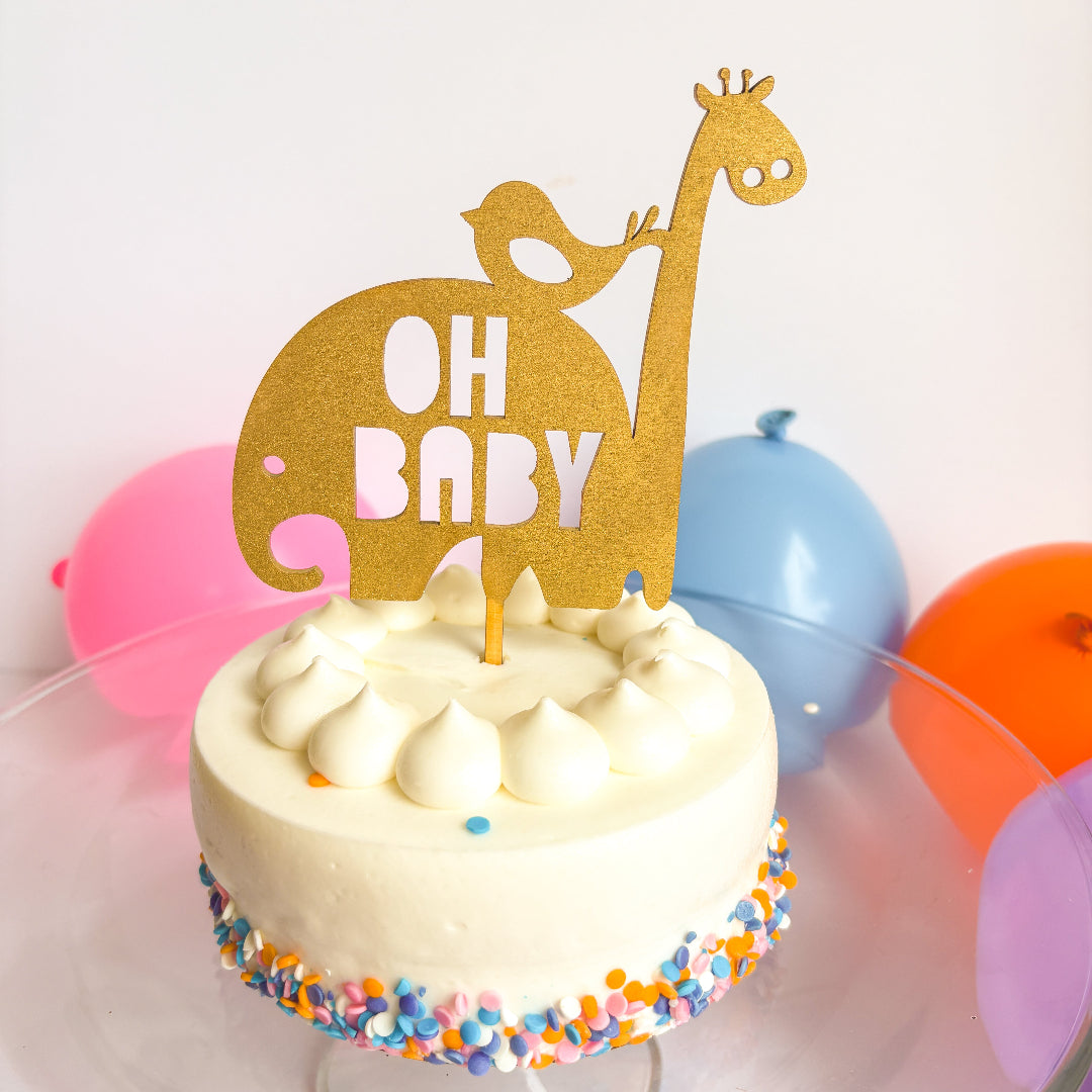 Small white cake with colorful sprinkles, a gold 'OH BABY' giraffe topper, and balloons in the background.