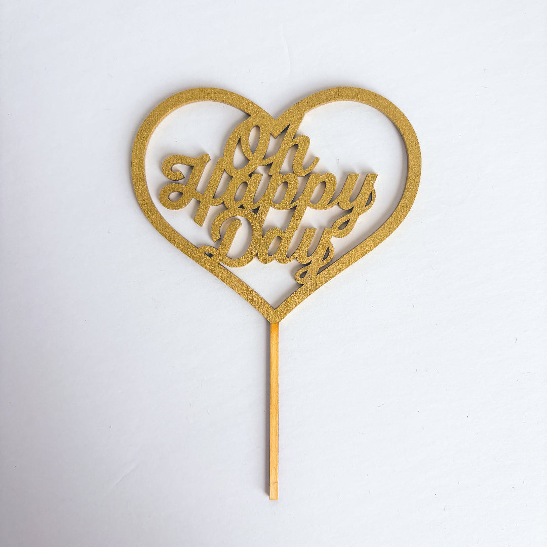 Gold heart-shaped cake topper with 'Oh Happy Days' text on a white background