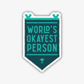 A humorous banner-shaped sticker with a dark teal background and a thick black outline, featuring the text "WORLD'S OKAYEST PERSON" in a lighter teal font. A small trophy icon is at the bottom of the banner.