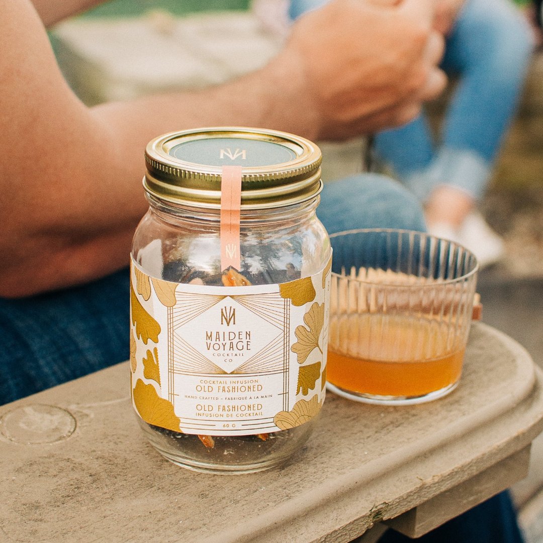 A glass jar filled with dried orange slices, dried cherries, and other ingredients. The jar has a white label with the Maiden Voyage logo and the product name "Old Fashioned Cocktail Infusion."