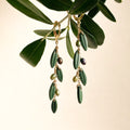 A pair of gold-tone drop earrings designed to look like olive branches, featuring dark green leaf-shaped drops, small olive-green beads, and dark purple seed beads, hanging from a real olive branch against a warm cream background.