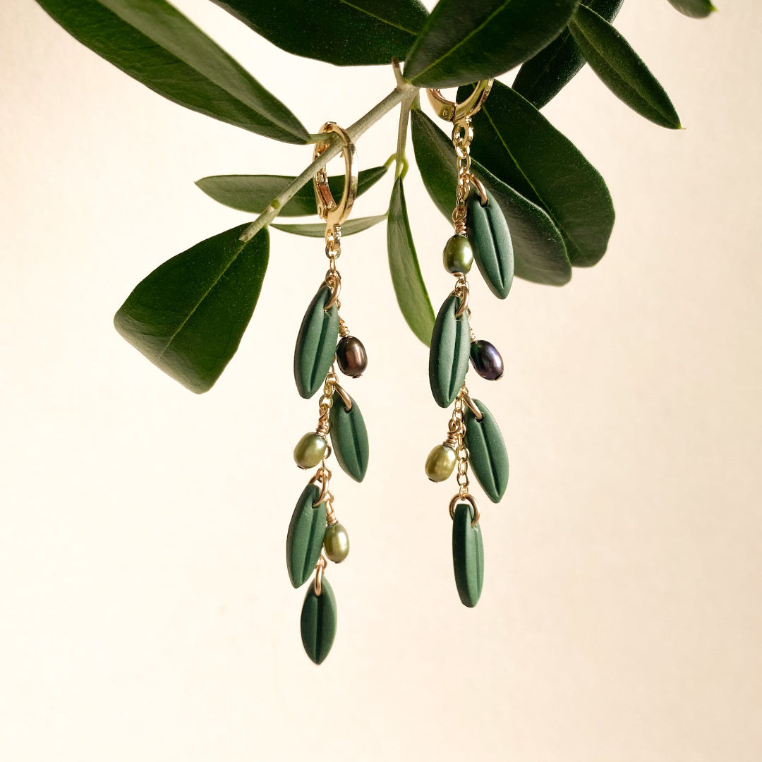 A pair of gold-tone drop earrings designed to look like olive branches, featuring dark green leaf-shaped drops, small olive-green beads, and dark purple seed beads, hanging from a real olive branch against a warm cream background.