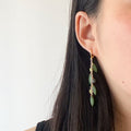 A close-up of a woman's ear wearing a long gold dangle earring with green bead-like drops, against a plain light background.