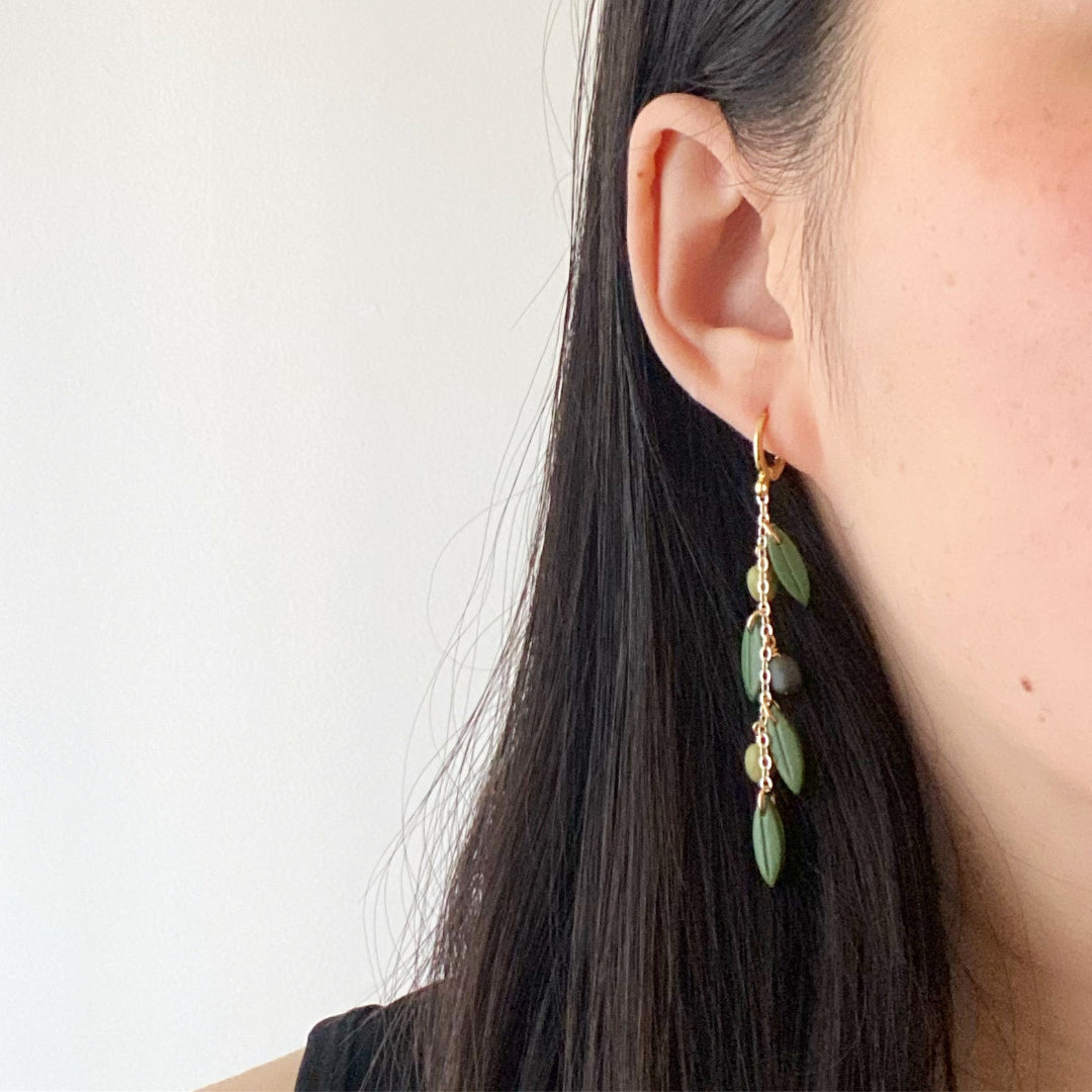 A close-up of a woman's ear wearing a long gold dangle earring with green bead-like drops, against a plain light background.