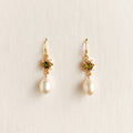 A pair of gold dangle earrings featuring a small green gemstone in a starburst setting and a dangling white pearl, shown on a plain off-white background.