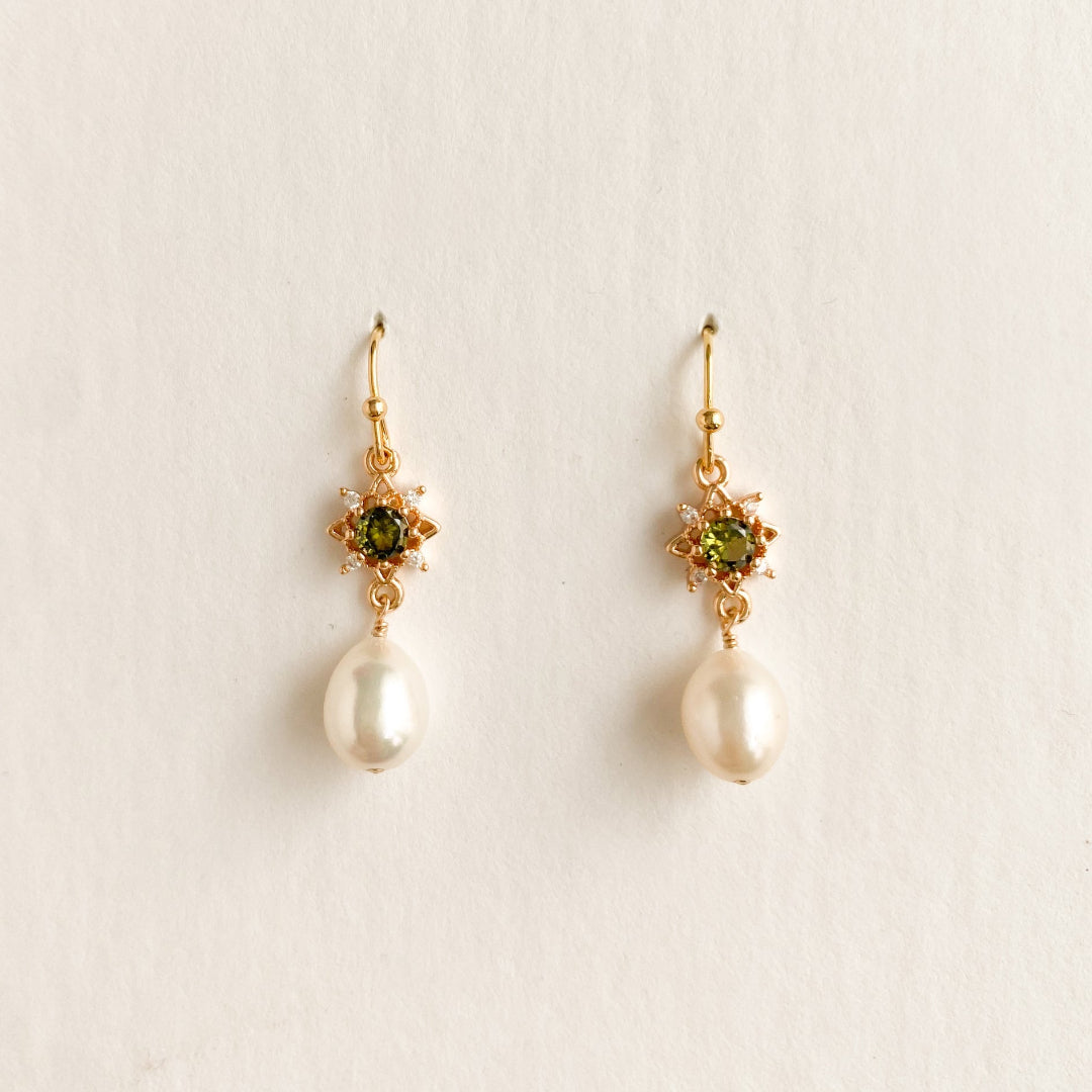 A pair of gold dangle earrings featuring a small green gemstone in a starburst setting and a dangling white pearl, shown on a plain off-white background.