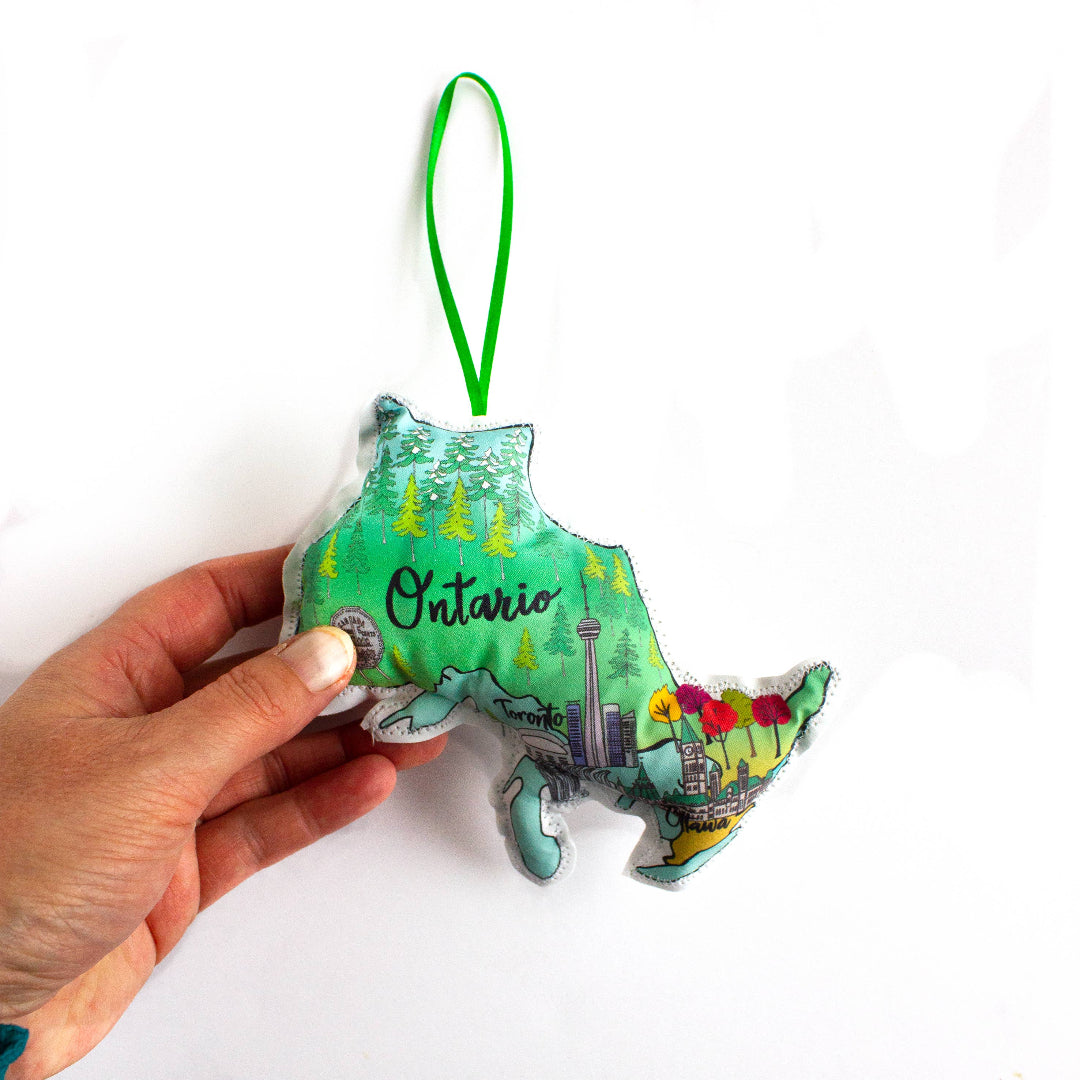 A close-up, high-angle flat lay shot on a plain white background features a soft, plush Christmas ornament shaped like the Canadian province of Ontario. The illustration on the ornament is divided into northern and southern regions, with the north showing dark green forests and the south showing the Toronto skyline and colorful autumn trees. The word "Ontario" is written in a dark script font in the center. A bright green ribbon loop is attached to the top for hanging.