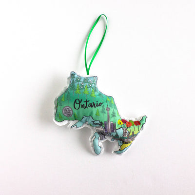 A close-up, high-angle flat lay shot on a plain white background features a soft, plush Christmas ornament shaped like the Canadian province of Ontario. The illustration on the ornament is divided into northern and southern regions, with the north showing dark green forests and the south showing the Toronto skyline and colorful autumn trees. The word 