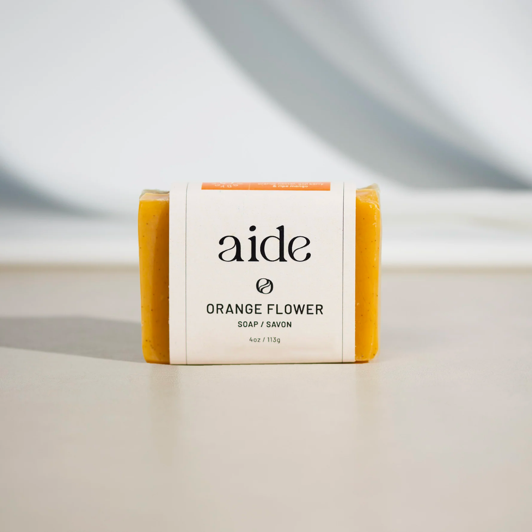 A bar of Aide Bodycare Orange Flower Soap, featuring a bright orange color and a paper label with the 'aide' logo and 'Orange Flower' text, displayed on a light-colored surface with a shadow overlay. The soap is natural and handmade, available in Ottawa, Ontario, Canada.