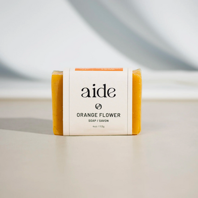 A bar of Aide Bodycare Orange Flower Soap, featuring a bright orange color and a paper label with the 'aide' logo and 'Orange Flower' text, displayed on a light-colored surface with a shadow overlay. The soap is natural and handmade, available in Ottawa, Ontario, Canada.