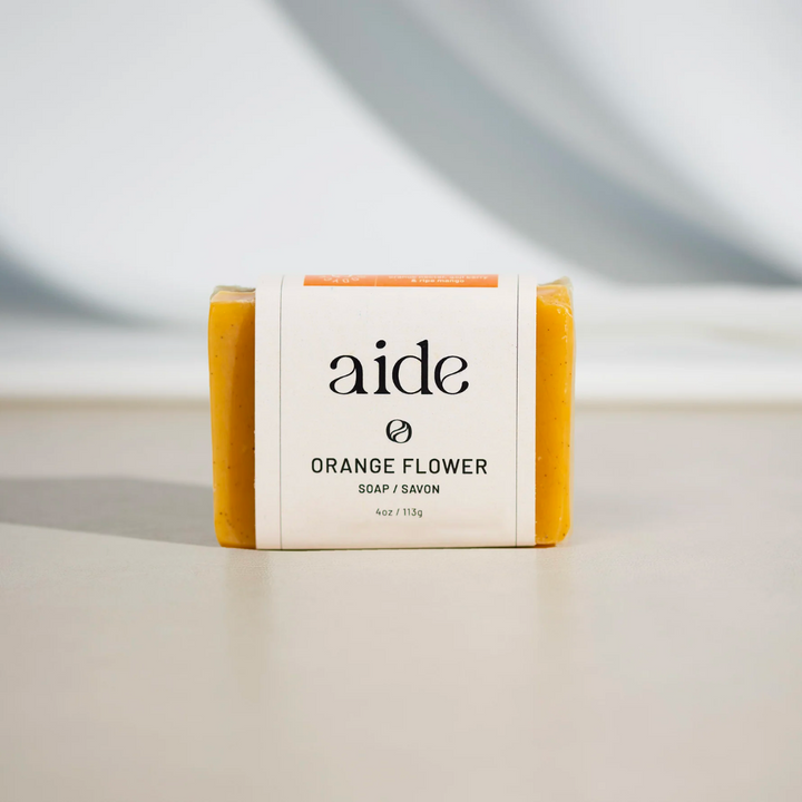 A bar of Aide Bodycare Orange Flower Soap, featuring a bright orange color and a paper label with the 'aide' logo and 'Orange Flower' text, displayed on a light-colored surface with a shadow overlay. The soap is natural and handmade, available in Ottawa, Ontario, Canada.