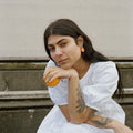 Woman in a white dress sitting on steps holding an orange, with a building in the background.