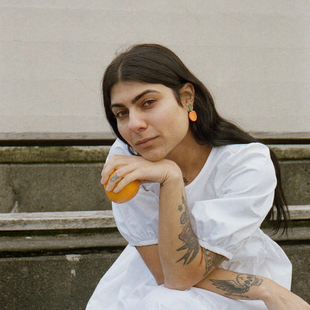 Woman in a white dress sitting on steps holding an orange, with a building in the background.