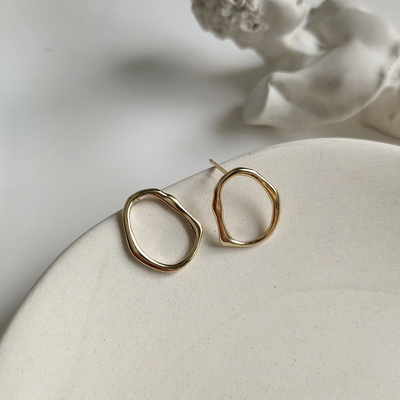 A pair of organic oval stud earrings made of gold fill, featuring a free-form, slightly irregular oval shape with a hammered texture, displayed on a textured ceramic surface. These modern earrings are available in Ottawa, Ontario, Canada.