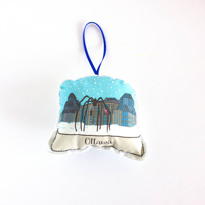 A close-up, high-angle flat lay shot on a plain white background features a soft, plush Christmas ornament shaped like a snow globe. The illustration inside the globe features the modern glass and stone architecture of the National Gallery of Canada in Ottawa, along with the large 