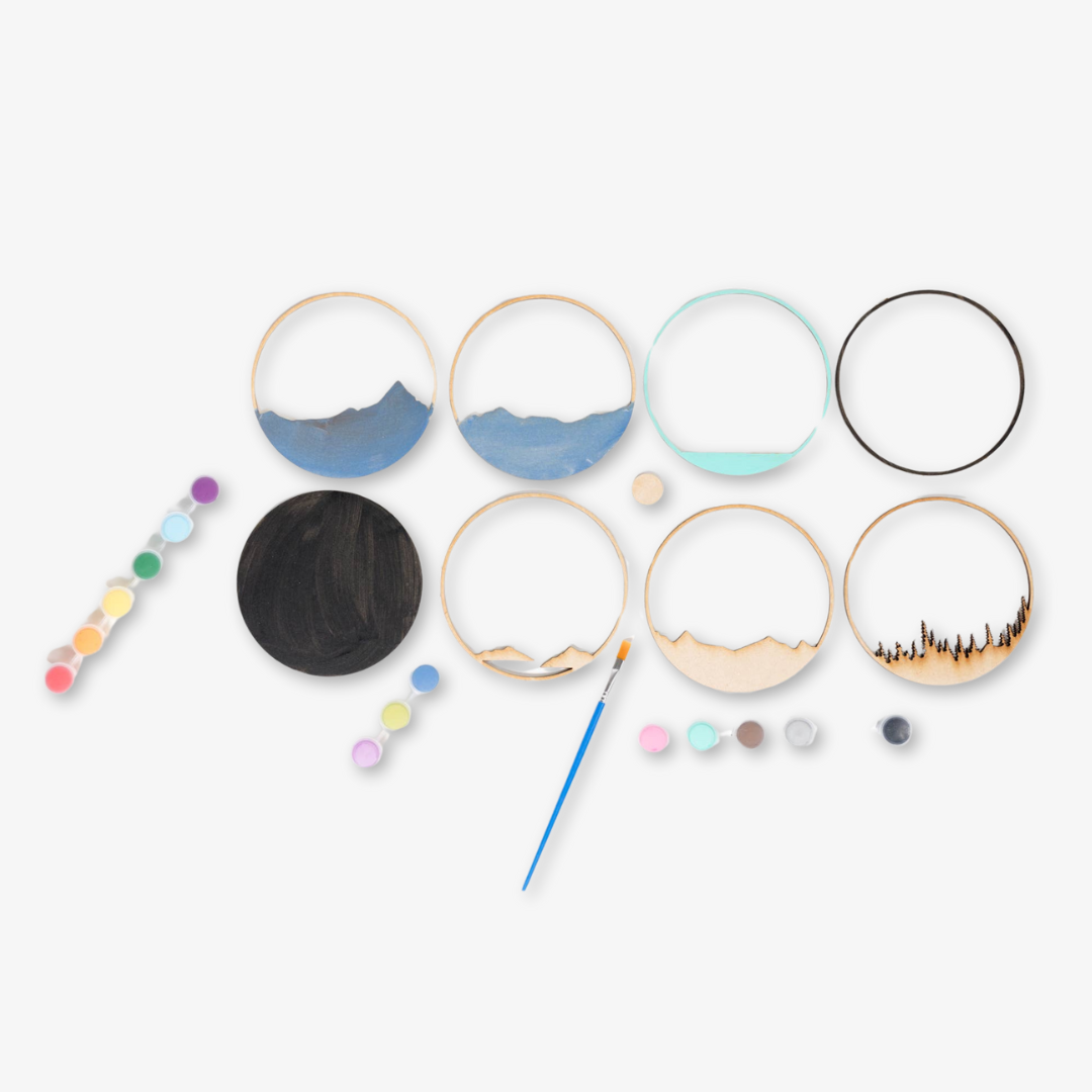 A Paint By Layers Craft Kit featuring a circular shadow box with a pre-designed landscape scene. The scene depicts a mountain range under a starry night sky, with a river flowing through a valley. The kit includes layers of wood, paint, and instructions. The title of the kit is "Rocky Mountain Shadow Box."