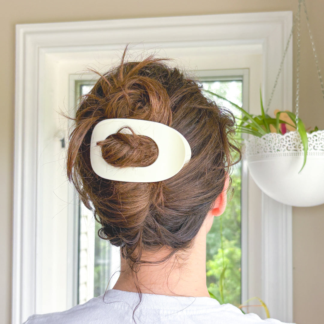 Person with a hair bun held in place by a large white hair clip, standing in a room with a window and plant.