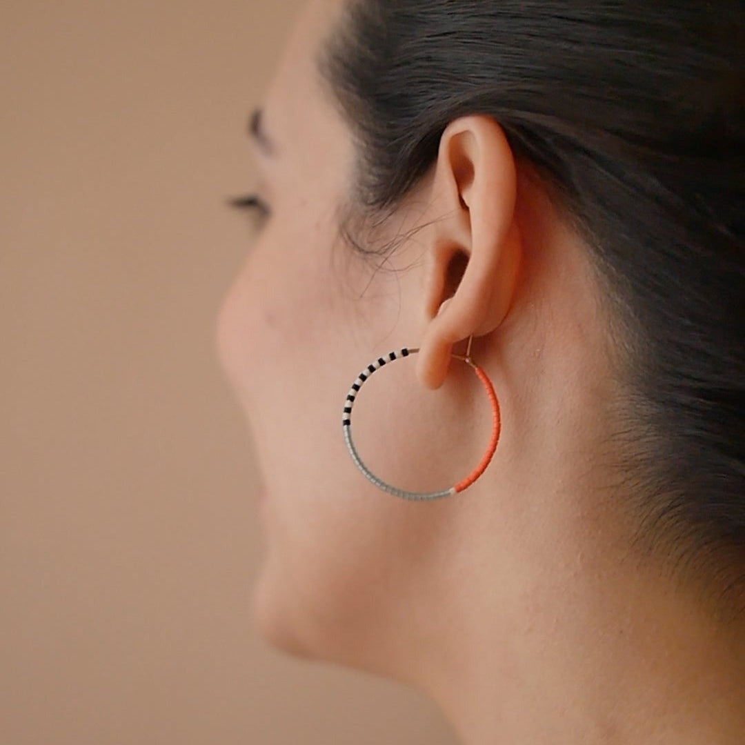A pair of delicate 14 KT gold hoop earrings, each partially threaded with small Japanese glass seed beads in a pattern of light blue, white, black, and coral. The beads cover approximately half of each hoop, leaving the remaining gold wire visible.