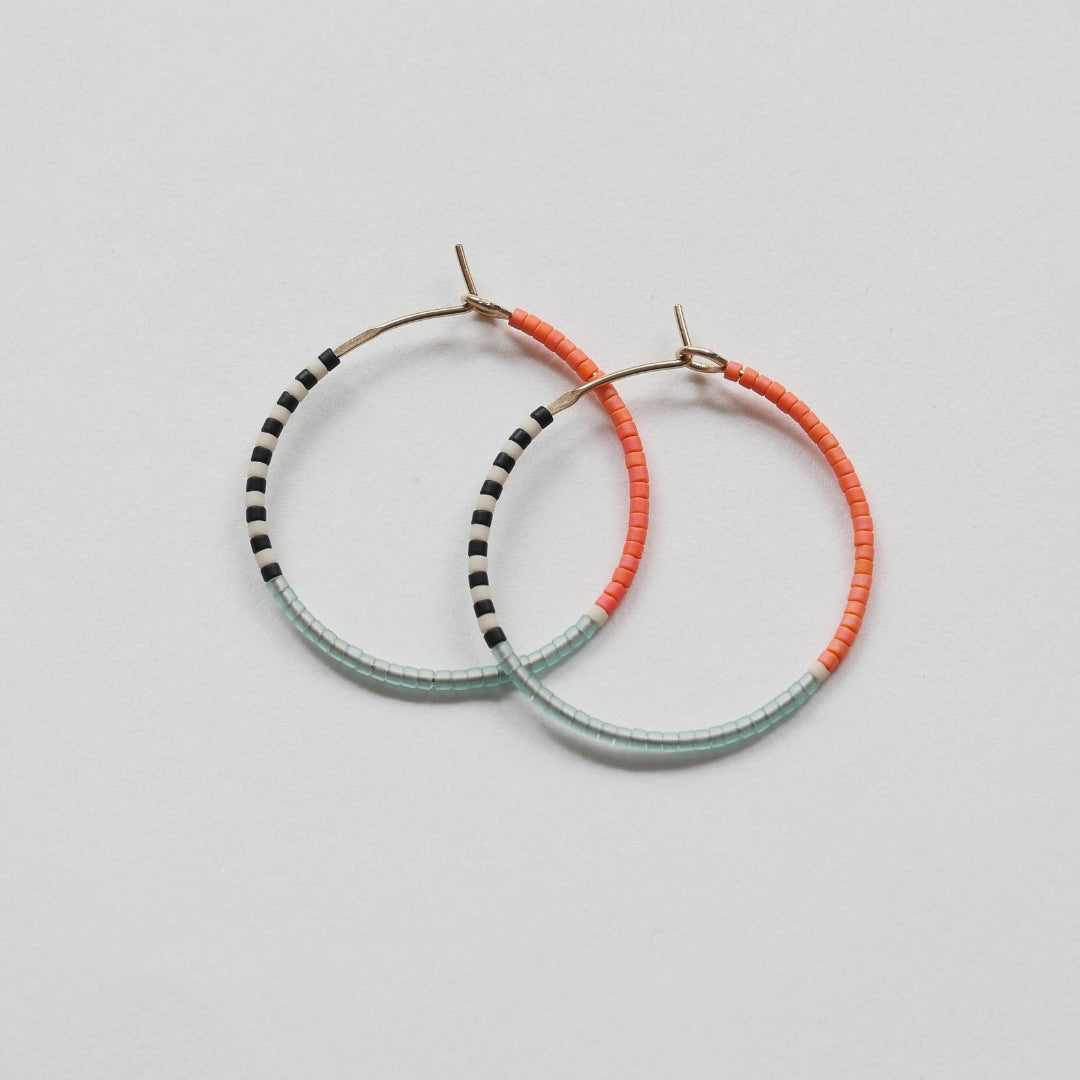 A pair of delicate 14 KT gold hoop earrings, each partially threaded with small Japanese glass seed beads in a pattern of light blue, white, black, and coral. The beads cover approximately half of each hoop, leaving the remaining gold wire visible.