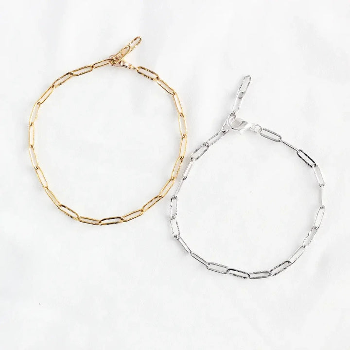 Two bracelets on a white surface. One bracelet is gold-colored, and the other is silver-colored. Both bracelets have a chain link design made of small, rectangular links.