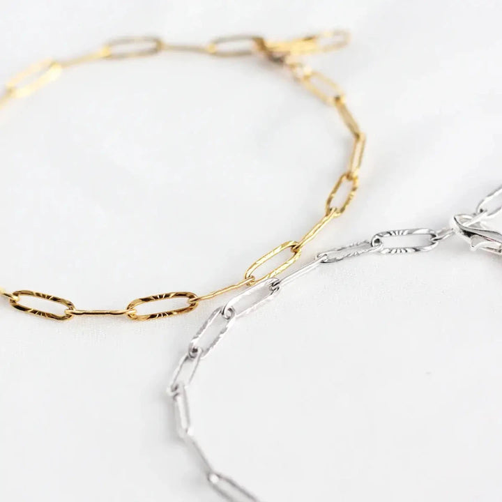 Two bracelets on a white surface. One bracelet is gold-colored, and the other is silver-colored. Both bracelets have a chain link design made of small, rectangular links.