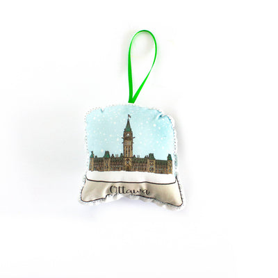 A close-up, high-angle flat lay shot on a plain white background features a soft, plush Christmas ornament shaped like a snow globe. The illustration inside the globe features the Peace Tower of the Canadian Parliament Buildings under a light blue, snowy sky. The base of the globe is white and says 