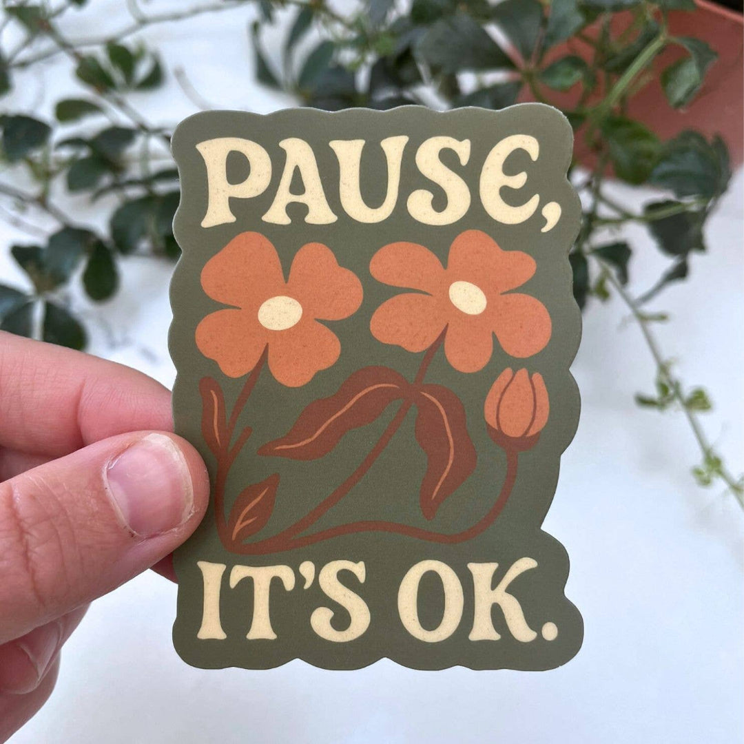 Sticker with flowers and text 'Pause, It's OK' held by a hand with a blurred plant background