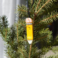 A yellow pencil-shaped Christmas ornament with a smiling face hangs from a black ribbon among green pine branches.